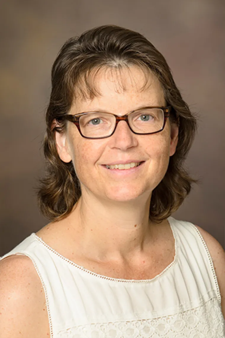 A professional headshot of a white woman and shoulder length brown hair, who is wearing classes and smiling at the camera